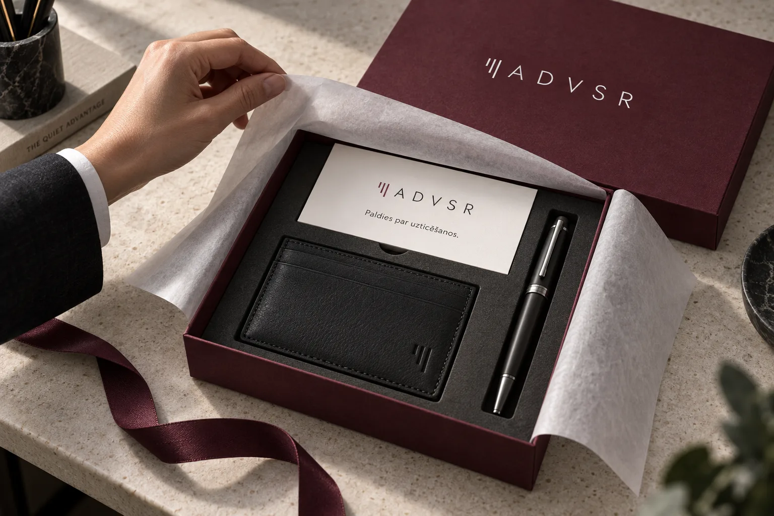 A hand opens a wine-coloured ADVSR gift box containing a card, a black accessory and a pen.