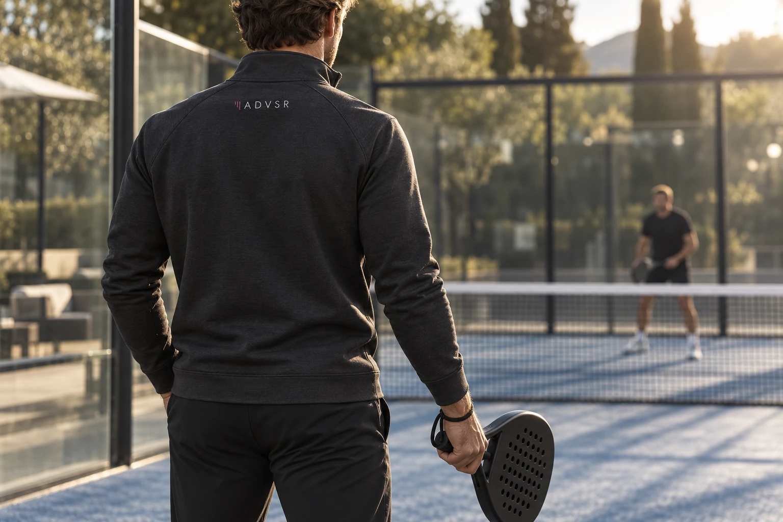 Rear view of a person holding a padel racket, wearing a top with a small ADVSR logo near the collar.